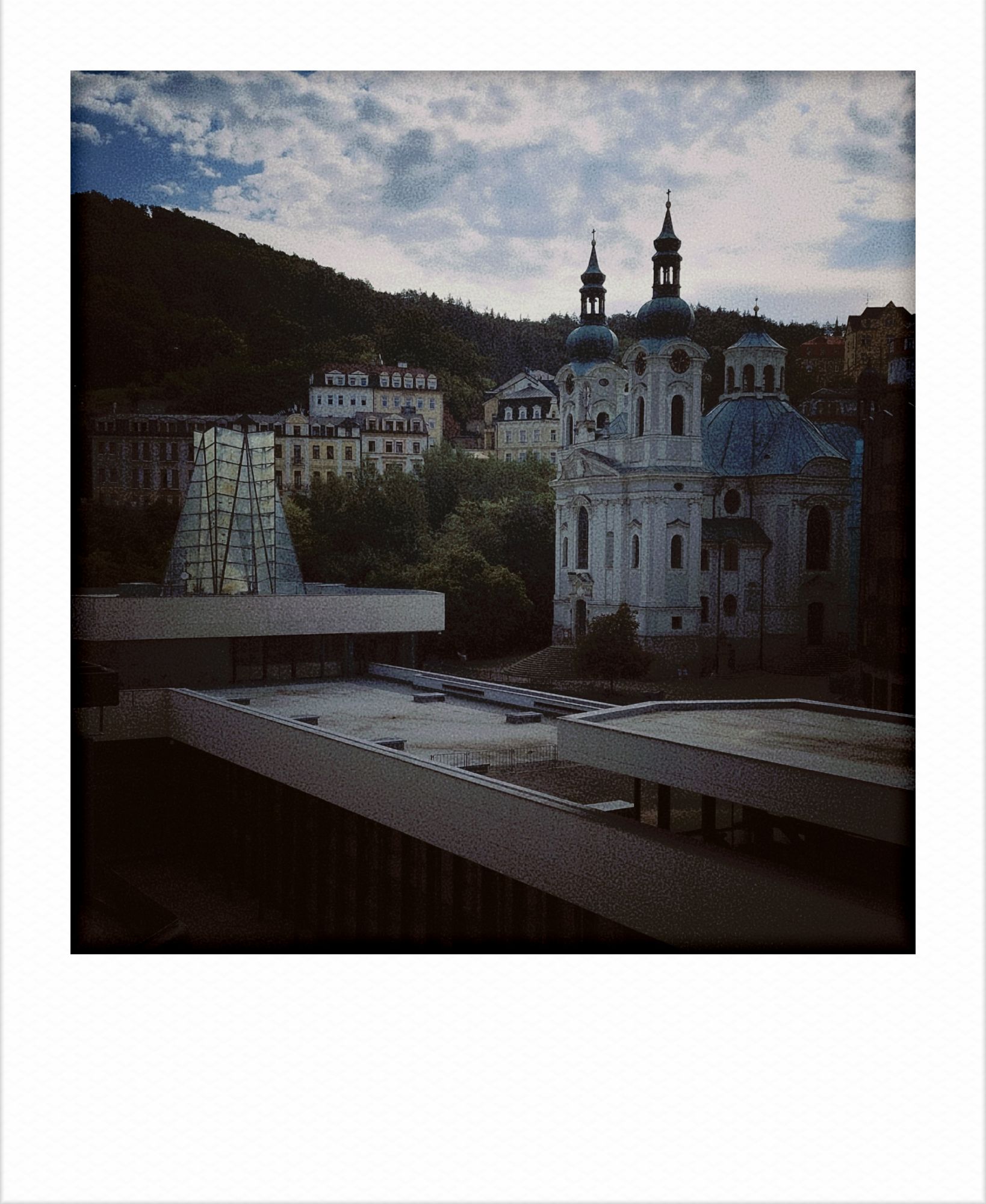 An East Modernist building, flat hall, in front, an old church in the right background surrounded by hills. Bright light, some clouds.